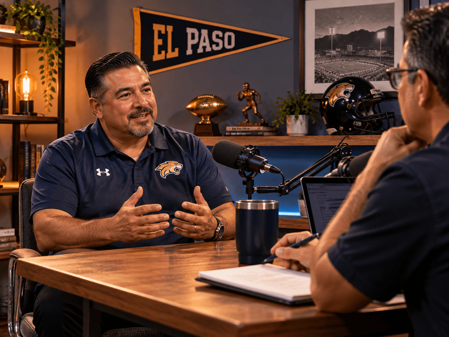 Athletic director in a polo shirt seated in a studio guest chair across a desk from a host with a microphone between them